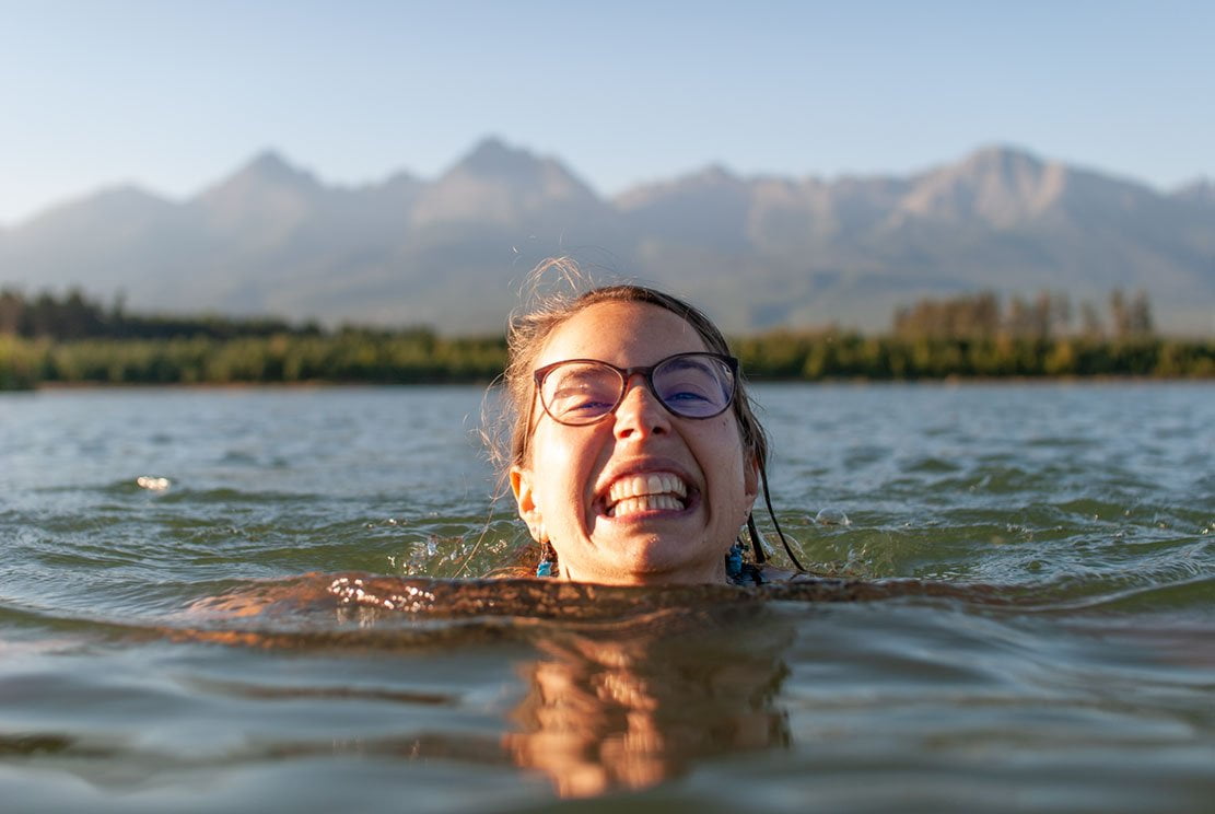 young beautiful woman smiling at you while swimming in a lake with mountains at the back