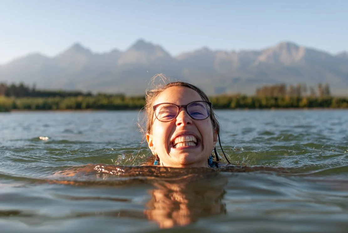 young beautiful woman smiling at you while swimming in a lake with mountains at the back
