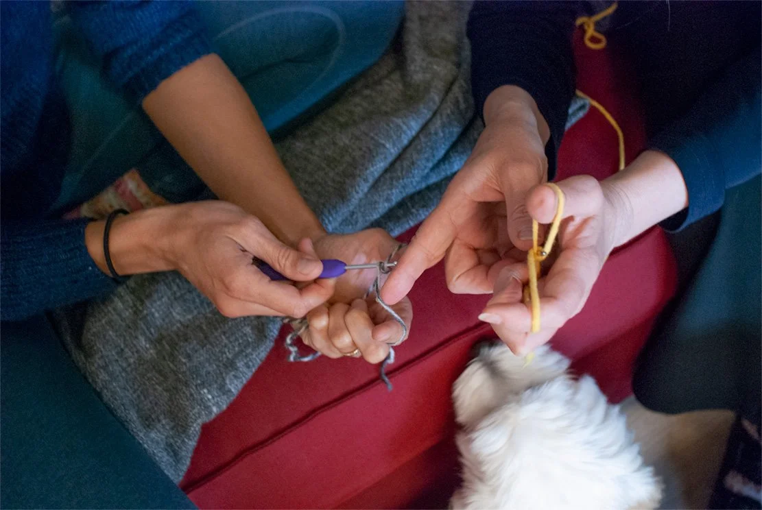 2 pairs of hands knitting where the pair on the right shows how to do it by pointing index finger to the other pair of hands knitting  