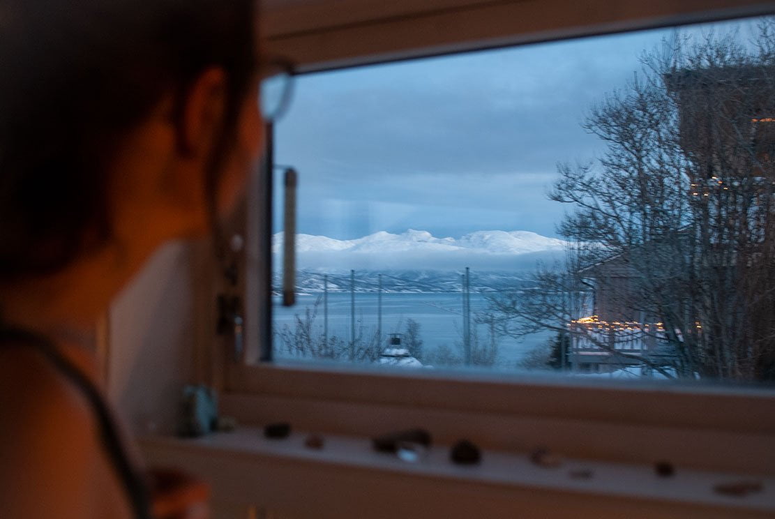 young woman looking out of a window towards snowed mountains on Ringvassøya island close by city of Tromsø