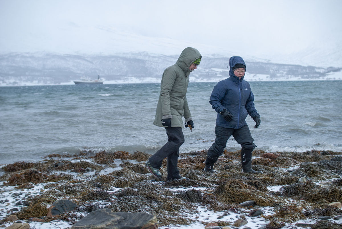 two women walking on a rocky beach in the Arctic circle surrounded by a fjord and snowy mountains on the other side